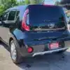 A 2018 Kia Soul EX in black is parked in a sunlit lot with a SUPER CLEAN dealership plate cover on the rear hatch. Other cars and trees are visible under a clear blue sky.