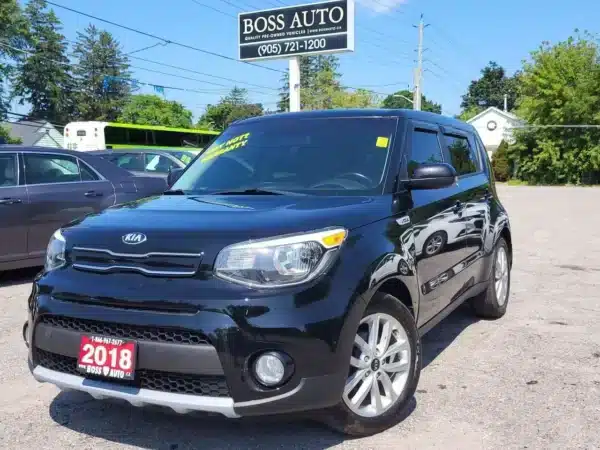 A 2018 Kia Soul EX is parked in a sunny lot, its 2018 license plate gleaming. With cars and trees in the backdrop, a Boss Auto sign stands prominently above, overseeing the scene.