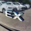 Close-up of a silver badge on a 2018 Kia Soul EX, highlighting its smooth, reflective surface. In the blurred background, a row of white cars is parked in a lot under sunny skies.