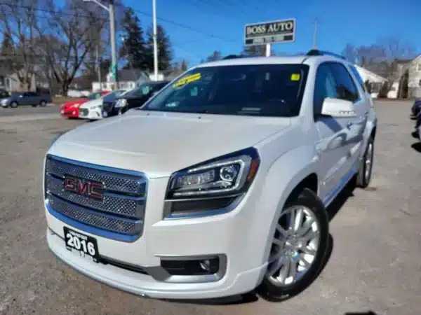 A white 2016 GMC Acadia Denali AWD is parked at a dealership with a yellow price sticker on its windshield. A BOSS AUTO sign stands in the background, surrounded by other cars, trees, and power lines.