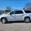 A 2016 GMC Acadia Denali AWD in white is parked on a gravel lot amidst other vehicles and trees under a clear blue sky.