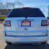 A white 2016 GMC Acadia Denali AWD SUV is parked on dirt under a clear blue sky. A red dealership plate reads LOADED with contact info, while sparse trees and a building provide background depth.