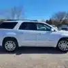 A 2016 GMC Acadia Denali AWD SUV sits on a gravel lot surrounded by other vehicles. Leafless trees and a clear blue sky emphasize its readiness for any adventure.