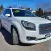 A 2016 GMC Acadia Denali AWD, marked by a Loaded sign, is parked on a paved lot amidst other cars. Its sleek white body stands out against the clear blue sky and bare trees.