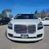 A 2016 GMC Acadia Denali AWD in white is parked at a dealership lot, featuring a For Sale sticker on its windshield amid signs and surrounded by other cars.