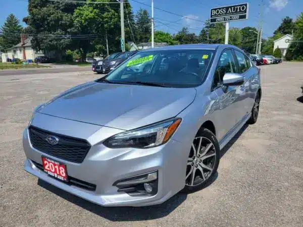 A 2018 Subaru Impreza Sport with Tech package in silver is parked at a car dealership, featuring a sunroof and sporty rims. A green sale sign is on the windshield, surrounded by trees and other cars, exemplifying its blend of sportiness and style.