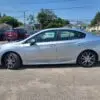 A 2018 Subaru Impreza Sport with a tech package, in silver, is parked on a gravel lot among other cars. Houses, trees, and power lines extend under the clear blue sky, crafting a sporty yet serene scene.