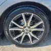 Close-up of a 2018 Subaru Impreza Sports front wheel, showcasing an alloy rim with a geometric design that enhances its sporty appeal. The black tire displays brand markings on the sidewall, while the cars body partially reflects sunlight on the asphalt below.