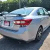 A silver 2018 Subaru Impreza Sport sedan with a spoiler and alloy wheels is parked in the lot. The Loaded dealership plate suggests it includes the Tech pkg. Other cars and trees are visible under a clear blue sky in the background.