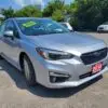 The 2018 Subaru Impreza Sport with tech package sits on a gravel lot with trees in the background. A yellow Certified sign is on the windshield, while a red 2018 plate is on the front bumper. Two other cars are faintly visible in the distance.