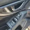 A close-up of the 2018 Subaru Impreza Sport w/ Tech pkgs interior door panel highlights a metallic handle, window and mirror control buttons, and a sport-inspired design featuring a tech package with a textured carbon-fiber-like finish.