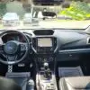 Interior view of a 2018 Subaru Impreza Sport w/ Tech pkg shows the dashboard, steering wheel, and gear shift. A yellow Sold sign is on the front windshield. Various controls and displays are visible with several cars parked outside.