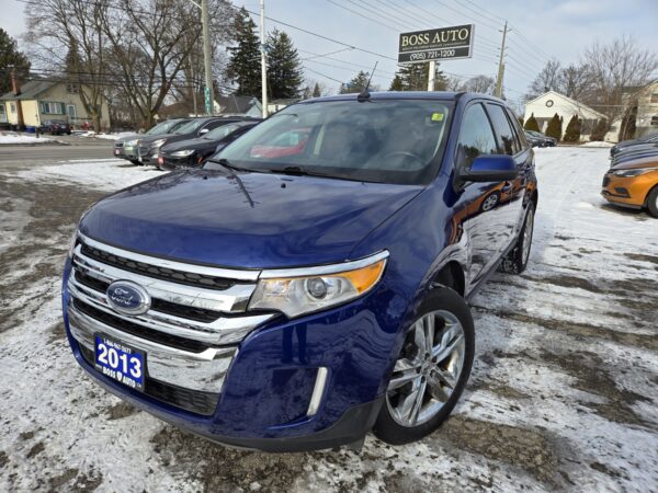 A blue 2013 Ford Edge SEL V6 SUV with chrome accents is parked on a snowy lot in front of Boss Auto, reflecting the wintry scene, while other cars and bare trees form the backdrop.