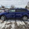 A 2013 Ford Edge SEL V6 in blue is parked on snow with other vehicles in the distance, surrounded by leafless trees and power lines under a partly cloudy sky, creating a perfect winter scene.