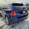 A 2013 Ford Edge SEL SUV, with a powerful V6 engine, is parked on a snowy surface by a road surrounded by bare trees under a cloudy sky, showcasing a Certified sign attached to its rear.