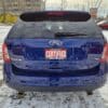 A 2013 Ford Edge SEL V6 in blue is parked in a snowy lot. The license plate features a Certified Boss Auto sign, with buildings and trees visible in the background.