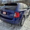 The 2013 Ford Edge SEL V6, painted blue, is parked on a snowy surface with a Certified dealer plate displayed at the rear. Snowy surroundings with trees frame the scene, while a red vehicle is partially visible on the left.