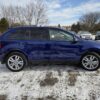 A 2013 Ford Edge SEL V6 SUV, in blue, is parked on a snowy lot. It is shown in profile view with its sleek design and shiny silver wheels. Other cars can be seen in the background alongside leafless trees under a partly cloudy sky.