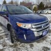 A blue 2013 Ford Edge SEL V6 SUV is parked in a snowy lot at a used car dealership, with the dealership sign and other cars visible in the background.