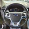 View of a 2013 Ford Edge SEL V6 interior from the drivers seat, highlighting the steering wheel with controls, speedometer, and dashboard display. Snow-covered ground and parked cars are visible outside this sleek marvel.