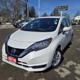 A white 2018 Nissan Versa Note is parked on an asphalt lot near a car dealership. The dealership sign reads Boss Auto. The sky is clear, and some trees and other cars are visible in the background.