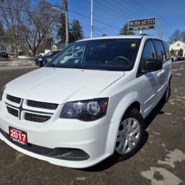 A white 2017 Dodge Grand Caravan SE is parked in a lot, showcasing its front and side. In the background, there are other vehicles, a Boss Auto dealership sign, and snow-dusted trees.