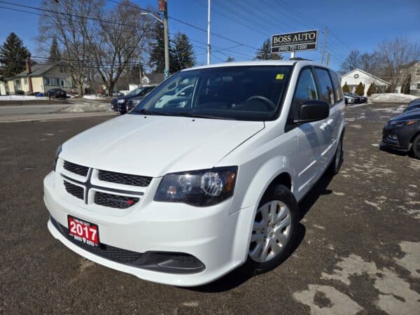 A white 2017 Dodge Grand Caravan SE is parked in a lot, showcasing its front and side. In the background, there are other vehicles, a Boss Auto dealership sign, and snow-dusted trees.