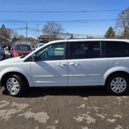 A 2017 Dodge Grand Caravan SE in white is parked in a lot on a sunny day, surrounded by other vehicles and trees, under a clear blue sky.