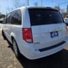 A white 2017 Dodge Grand Caravan SE, parked in a sunlit outdoor lot, has the rear license plate Like New. Snow and vehicles can be seen amidst trees and power lines under the clear blue sky.