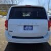 The image shows the rear of a white 2017 Dodge Grand Caravan SE, parked outdoors with a LIKE NEW license plate displaying BOSS AUTO and a phone number. Other vehicles and bare trees are in the background.