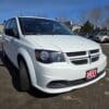 A white 2017 Dodge Grand Caravan SE with a front license plate parked on pavement, surrounded by trees and other parked cars under a clear blue sky.