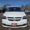 A front view of a 2017 Dodge Grand Caravan SE, white minivan, sits in a car lot with a Boss Auto license plate. Other vehicles can be seen behind it under a clear blue sky.