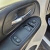 Close-up of a 2017 Dodge Grand Caravan SEs interior door panel featuring a chrome handle and power window controls for adjustments and locking. Sunlight creates shadows on the panel, with the floormat suggesting this minivans practical design.