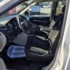 Interior of a 2017 Dodge Grand Caravan SE reveals dark fabric-upholstered driver and passenger seats, with the dashboard, steering wheel, and gear shift in view. A paper mat sits on the passenger floor as sunlight streams through the open door.