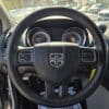 Close-up of the 2017 Dodge Grand Caravan SE steering wheel with the logo, set against a dashboard displaying speed and RPM gauges. Sunlight streams through the windows, creating shadows inside, while a snowy lot with a vehicle is visible outside.