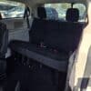 Inside the 2017 Dodge Grand Caravan SE, theres third-row seating with three upright black fabric seats and seatbelts, offering comfort. The rear window provides a view of parked cars outside.