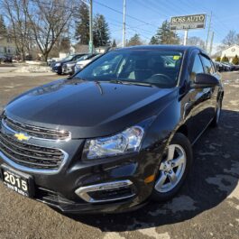 A black 2015 Chevrolet sedan is parked in a car lot outside a dealership. The foreground features the cars front and side, while other vehicles and a building are visible in the background on a clear day.