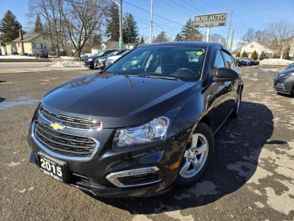 A black 2015 Chevrolet sedan is parked in a car lot outside a dealership. The foreground features the cars front and side, while other vehicles and a building are visible in the background on a clear day.