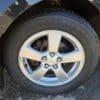 Close-up of a cars front wheel showing a silver alloy rim with a black Firestone tire. The hubcap features five bolts. The cars black body and part of the road surface are visible in the background.