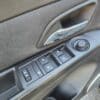 Close-up of a car door interior showing a silver handle and controls. The panel includes four window buttons and a circular knob, likely for adjusting mirrors. The door panel is upholstered in dark textured material.