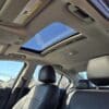 Interior view of a car showing black leather seats and an open sunroof. The sky is visible through the sunroof, and sunlight illuminates the interior. Trees and power lines are seen through the windows.