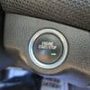 Close-up of a cars engine start/stop button surrounded by textured black material. The button has a silver rim and black center with ENGINE START/STOP text in white, and a small green line beneath.