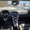 Interior view of a Chevrolet car showing the steering wheel, dashboard with a digital display, and black leather seats. Sunlight illuminates the showroom through the windshield. Floor mats say YOUR BUSINESS IS APPRECIATED. Snow is visible outside.