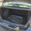 Open trunk of a black car, showing a spacious interior. Inside the trunk are a few stacked black floor mats. The car is parked in a sunlit area with visible pavement.