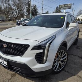 A white 2020 Cadillac XT4 SUV is parked in a dealership lot on a sunny day. The car features a prominent grille and sleek headlights. In the background, there are trees, a house, and a sign for BOSS AUTO.